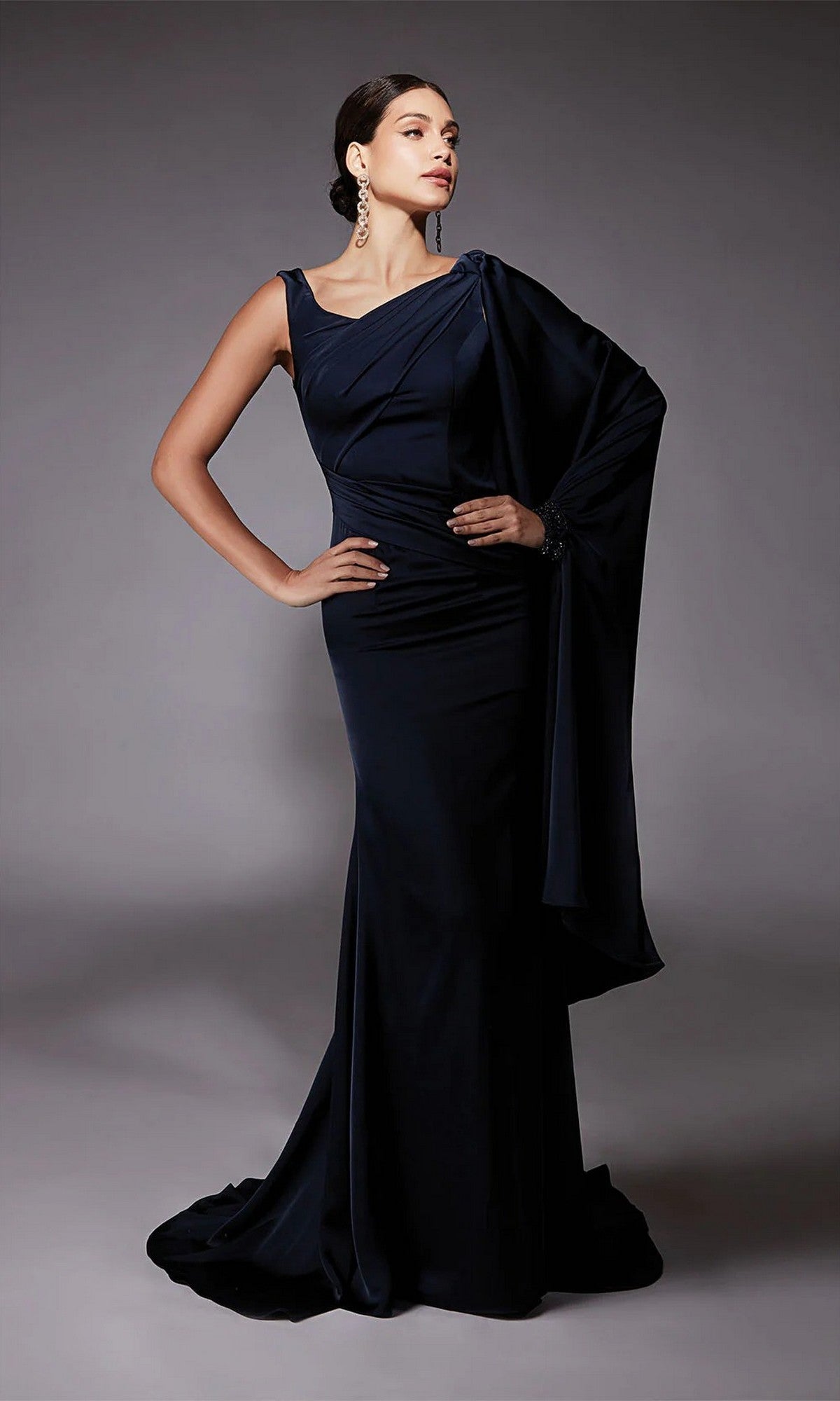 A woman models the Alyce 27722 Long Formal Dress—a floor-length, dark navy one-shoulder gown with a draped sleeve—posing with one hand on her hip against a plain gray background.